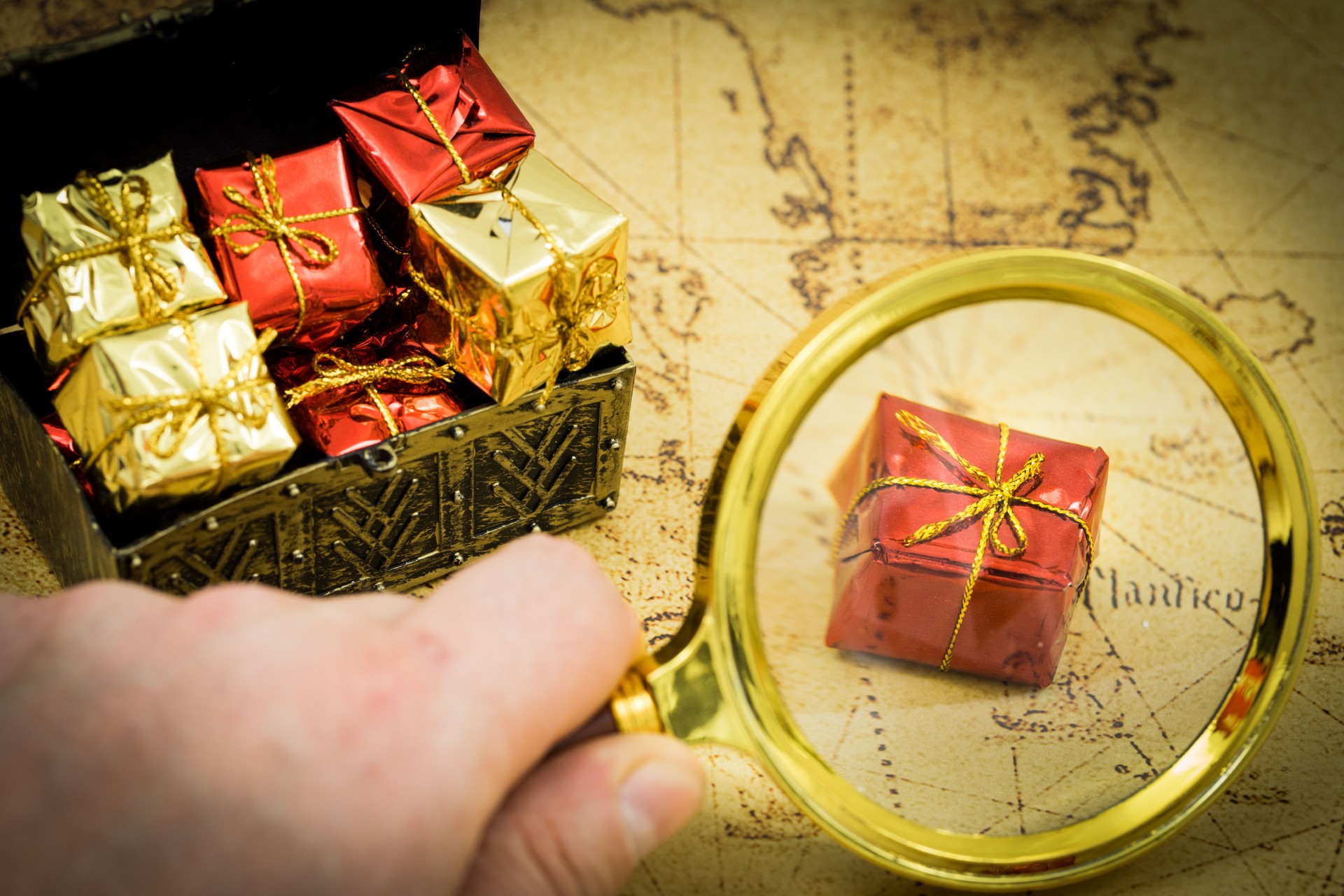 the treasure hunt. A hand with a magnifying glass above a chest full of valuables and gifts, In the background an old map for adventure hunters